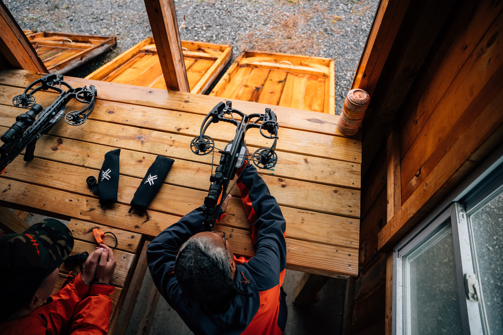 an aerial view of a person holding a crossbow sitting at a wooden table pointing it out a window