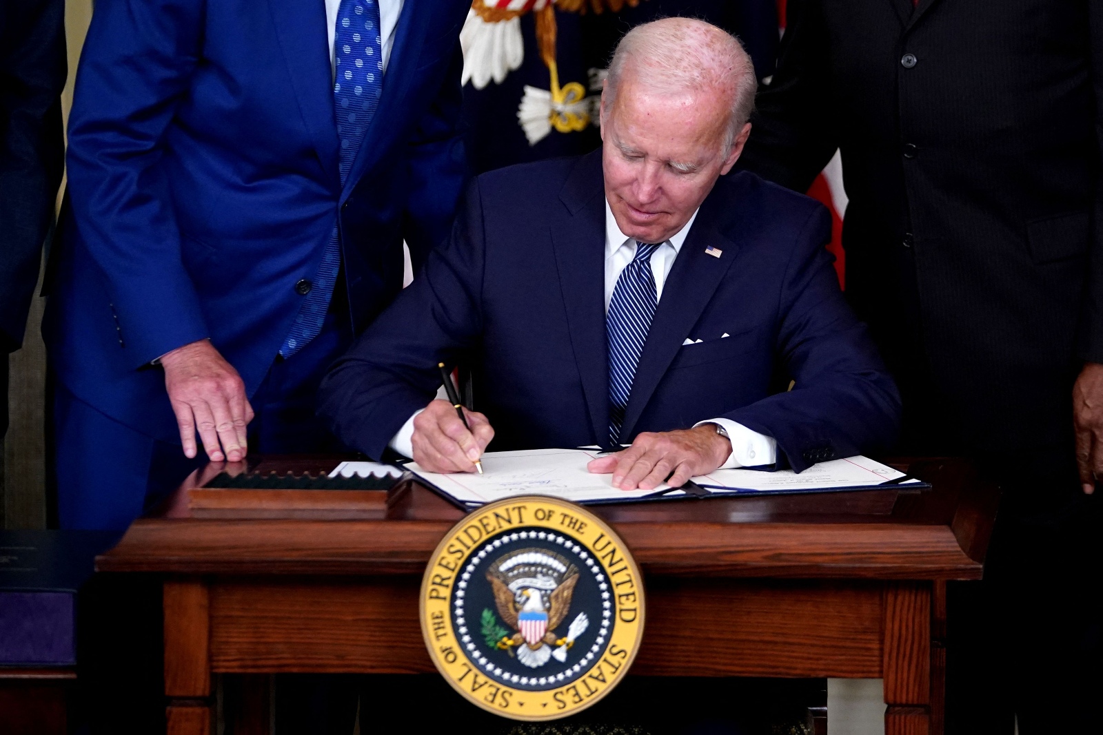 Biden signs a piece of paper as people in suits crowd around.