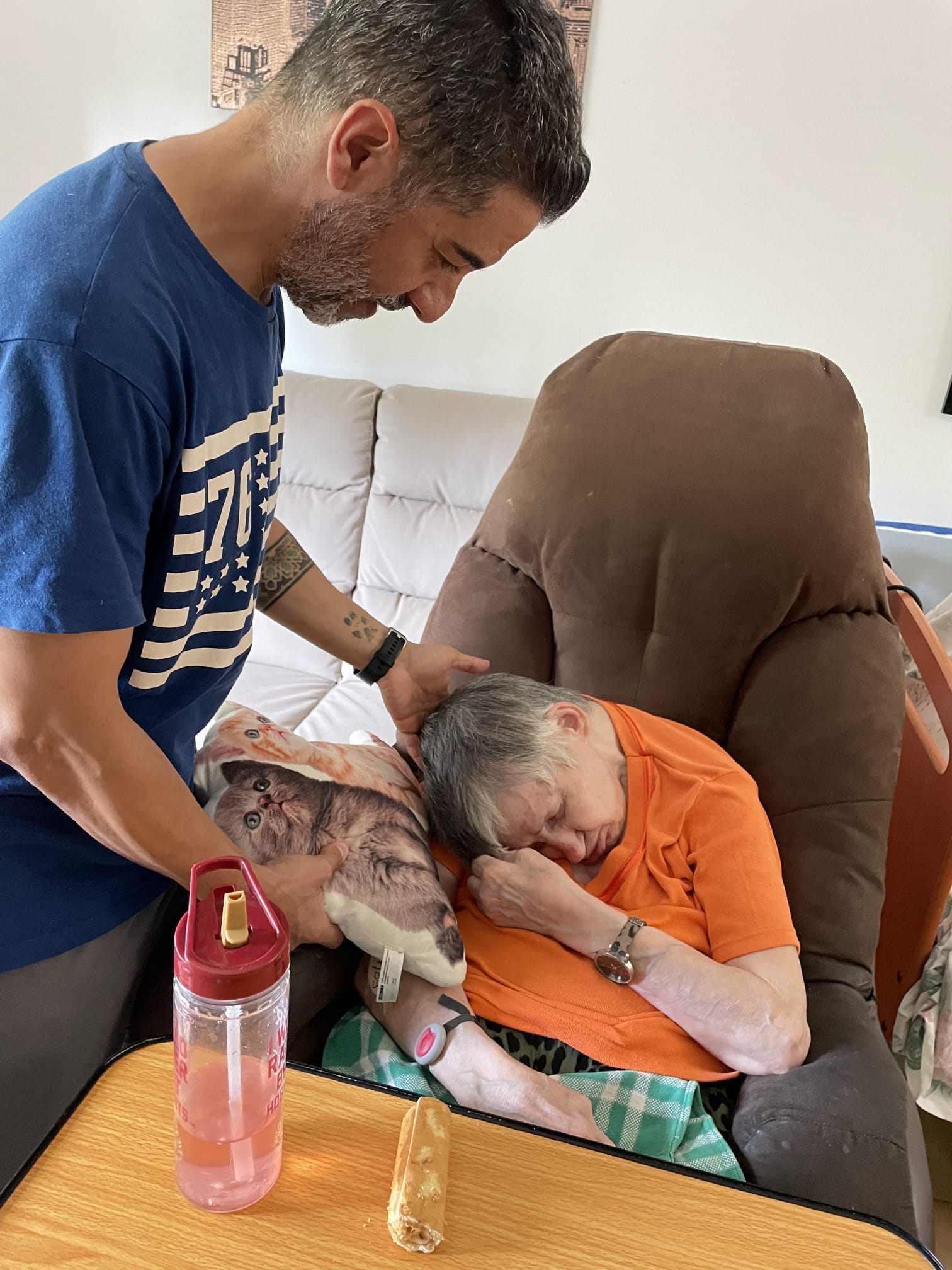 a man in a t-shirt puts a hand on an elderly woman's head as she sits in a chair