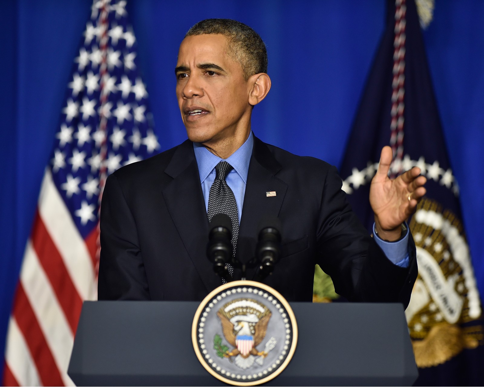 a man (Barack Obama) in a suit stands in front of an American flag and behind a podium