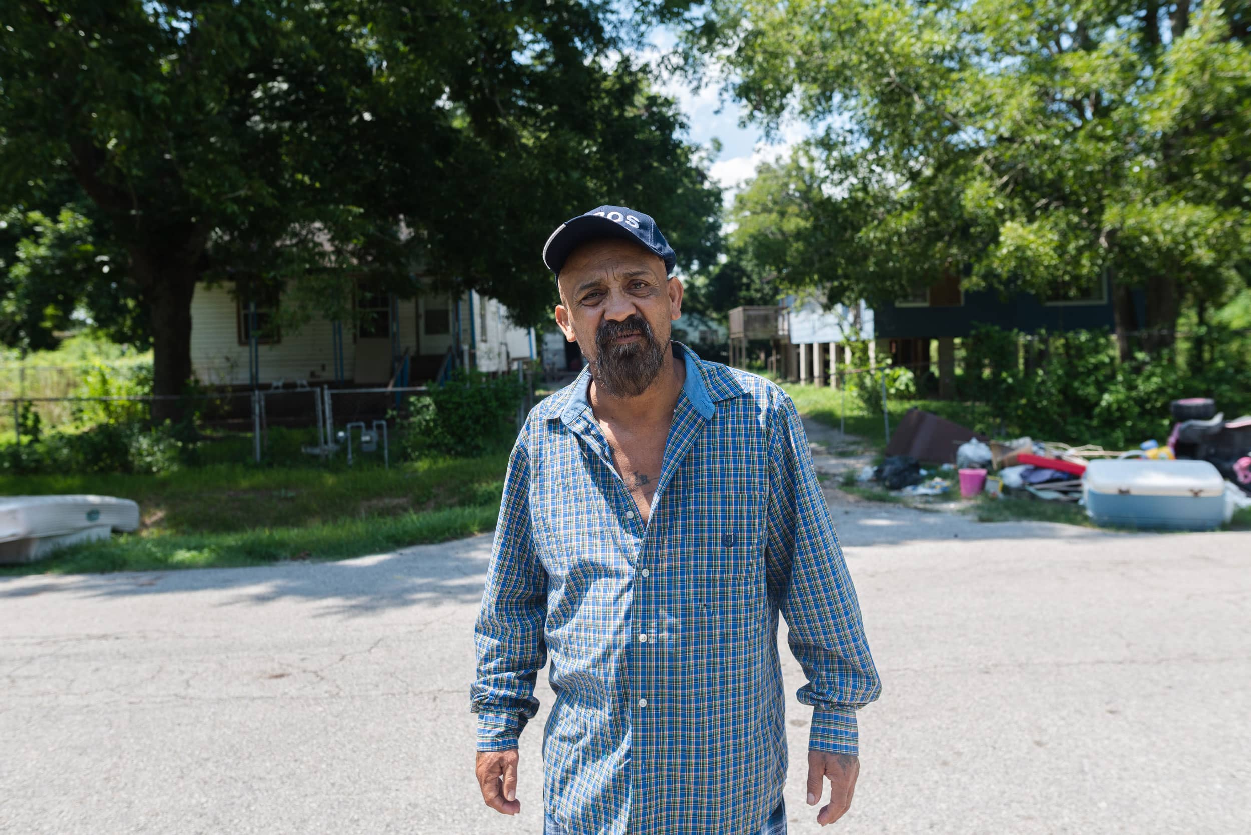 A man in a button up shirt stands in front of a rural house