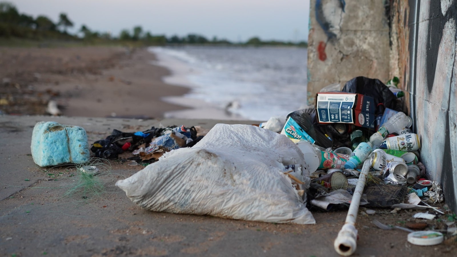 a plastic trash bag with lots of beer cans near a brick wall with sand in the background