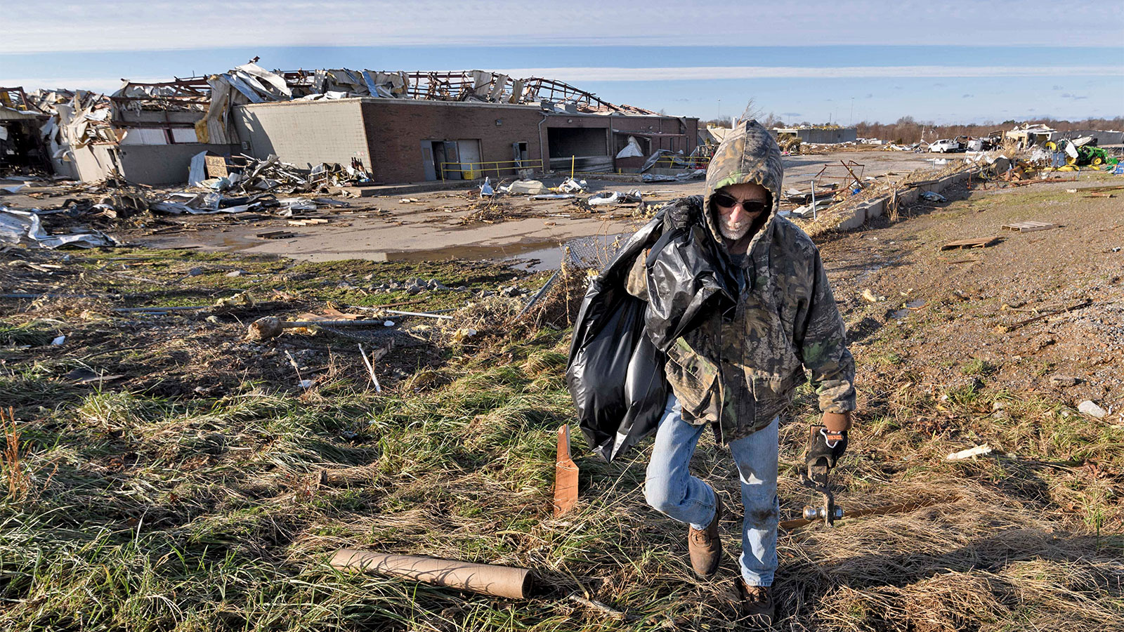 Man holding garbage bag walking towards camera with warehouse ruins behind him