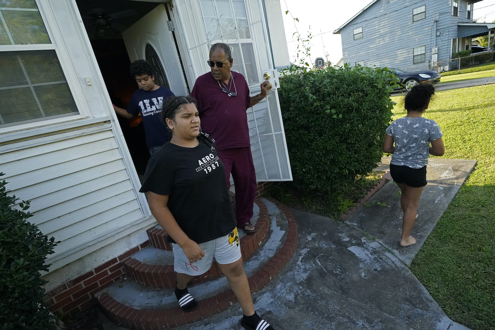 a man in a red shirt and red pants holds open a door through which three kids are walking