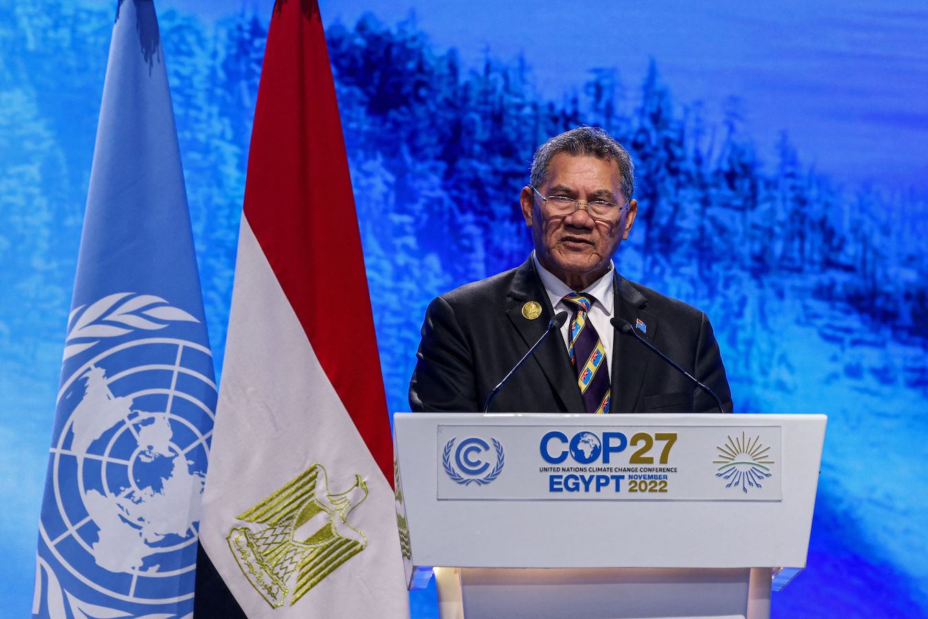 Man in a suit and tie stands at a podium in front of two flags