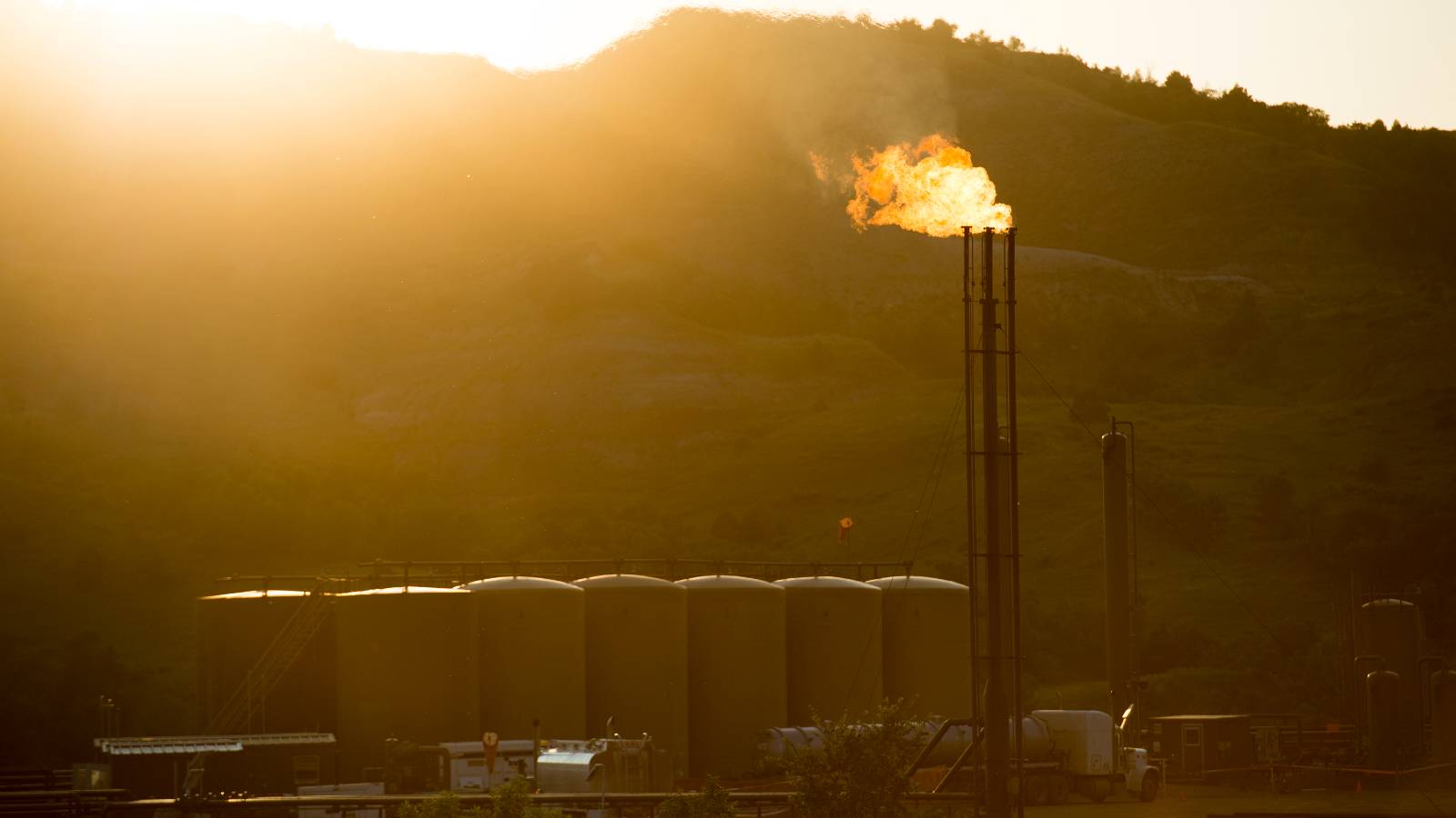 A gas well sits on the Fort Berthold Indian Reservation as the sun sets behind a ridge line. A tall metal structure is 