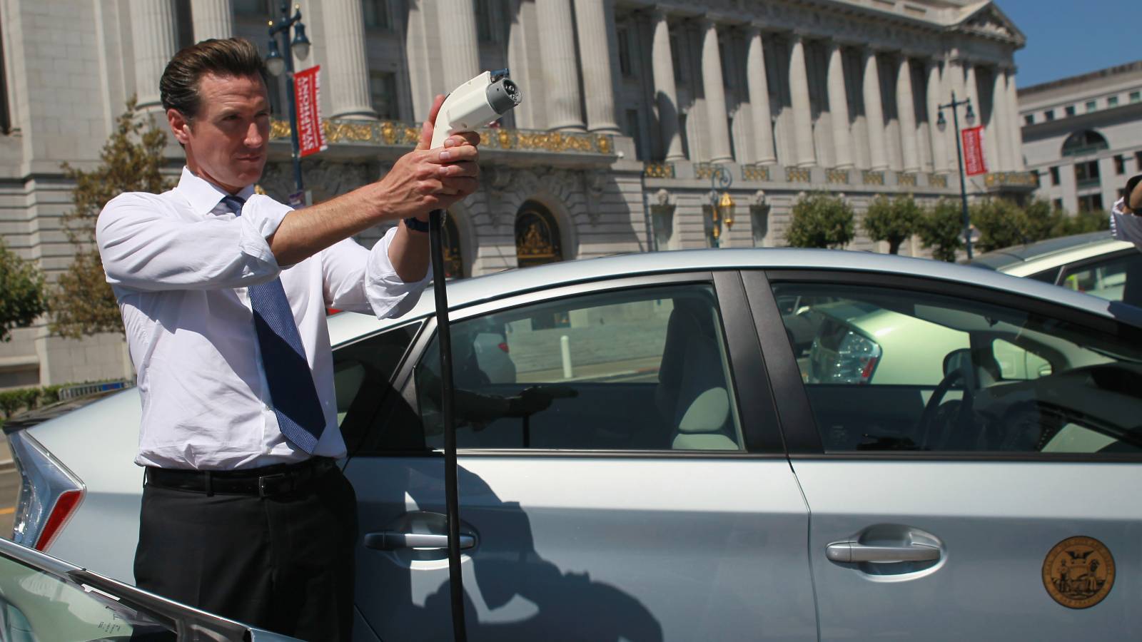 Governor Gavin Newsom stands next to a car holding an electric charger pointing it as though it were a gun