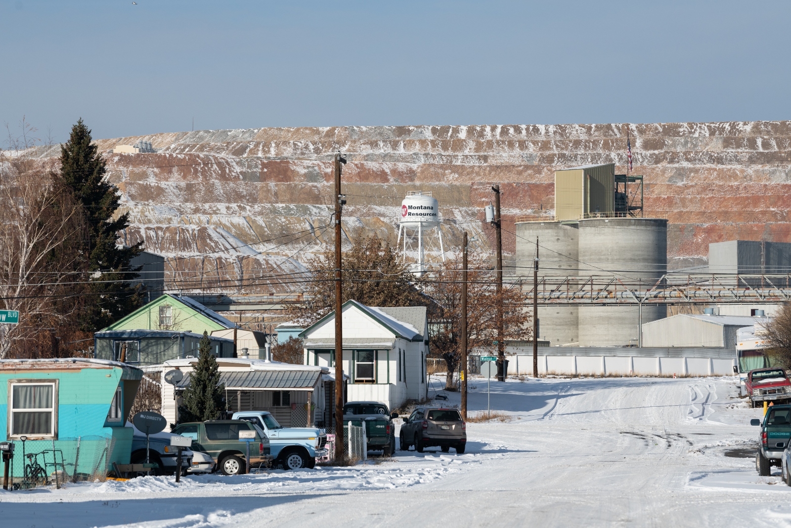 A residential neighborhood with two small houses and a blue truck sit on a snow-covered road in front of a wall of reddish earth, with industrial buildings in the foreground.