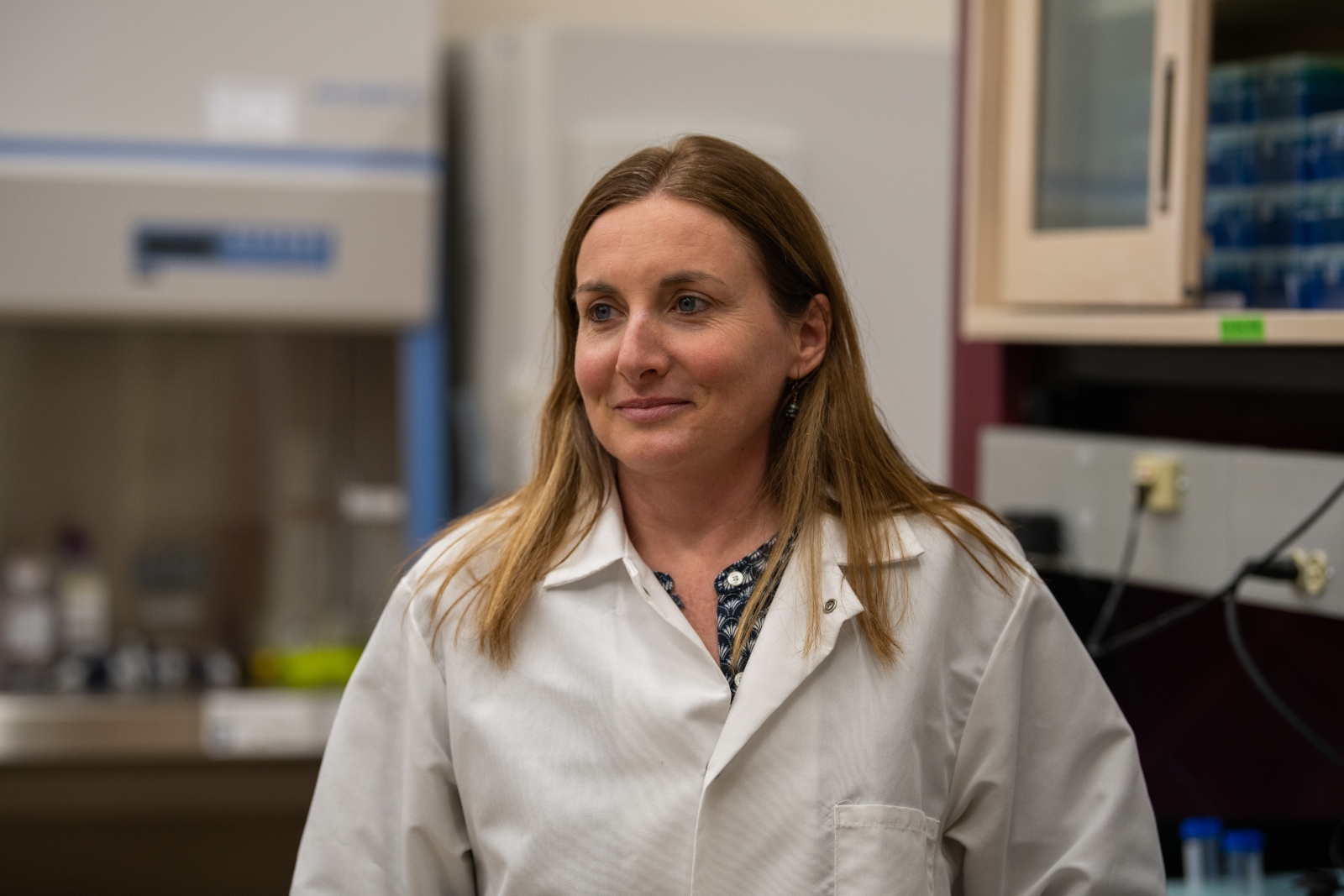 A blonde woman in a white lab coat is smiling.