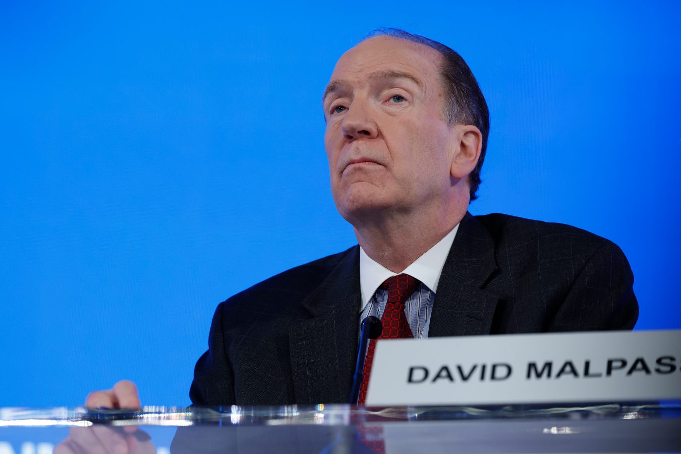 An older man in a dark suit sits at a podium with his name plaque saying 