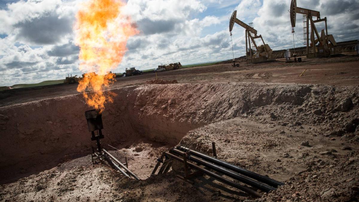 Gas pipes come out through the walls of a ditch and a bright orange flame vents out natural gas. Drilling equipment sits in the background.