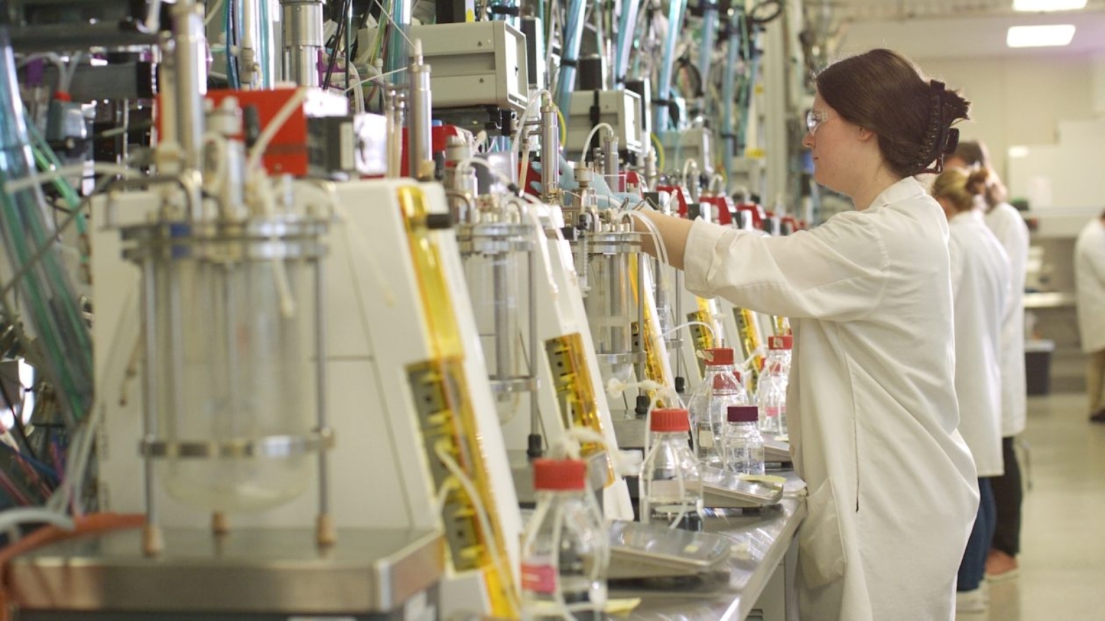 A scientist in googles and a white lab coat works in a row of white and yellow machines with lots of tubes.