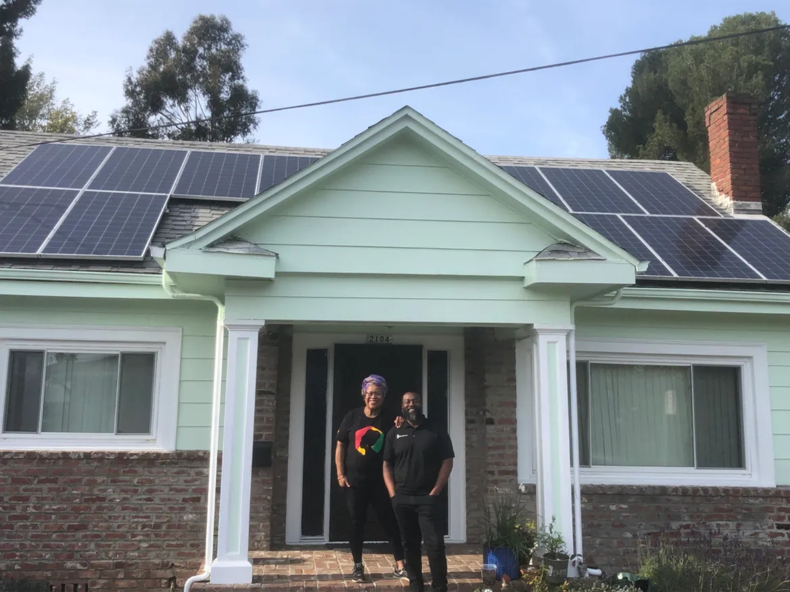 A man and a woman are smiling, and stand in front of a green house, with the roof covered in solar panels.