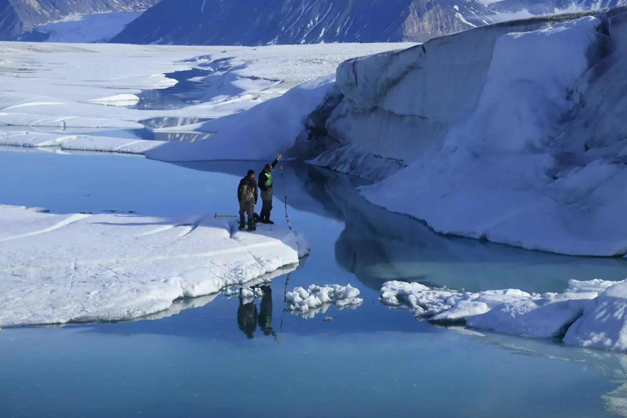 two figures dressed for cold weather look down into a stream of water between large ice flows