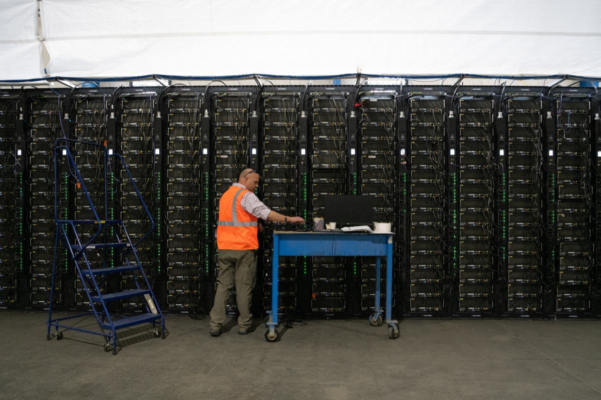 A man in a bright orange vest stands next to a blue cart in front of a huge row of black machines.