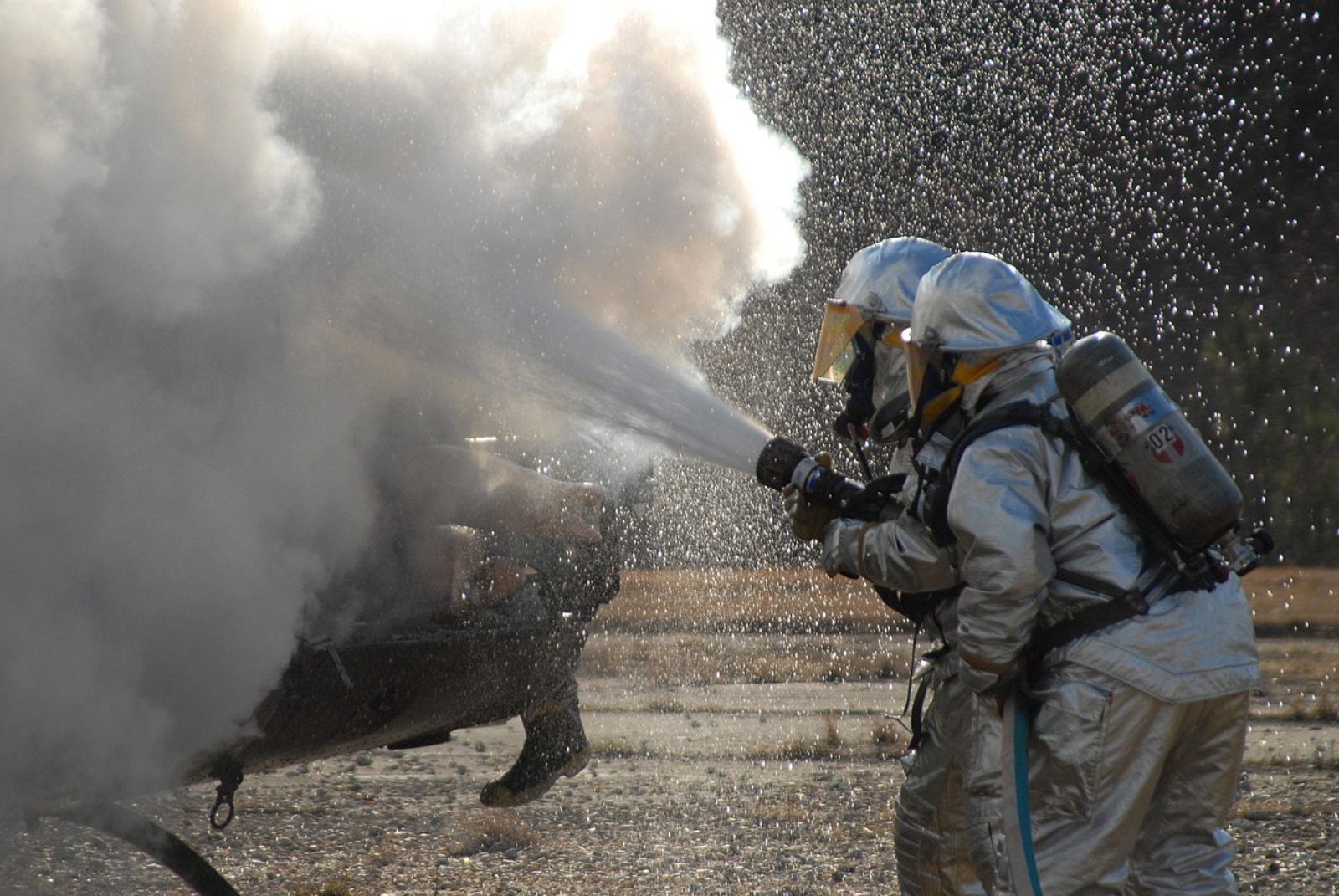 Two firefighters in silver protective gear spray white foam to put out a fire.