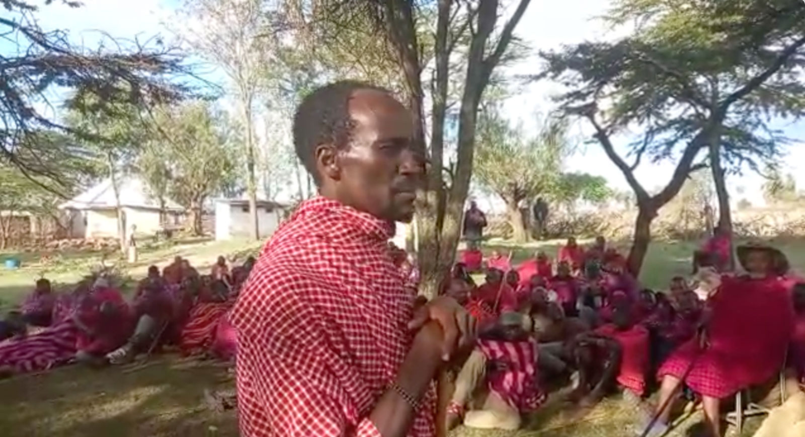 A man wearing a red and white blanket looks forward while other people also wearing blankets sit in the background