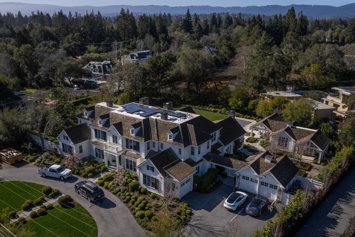 An aerial view of a mansion surrounded by trees with cars parked in front of the garage and street in front of the house.