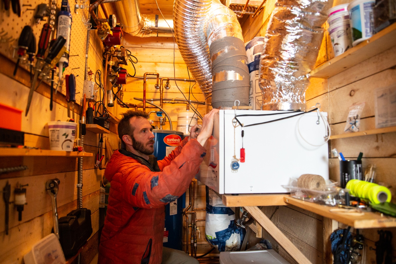 A bearded man in a red coat holds a large white box surrounded by pipes.