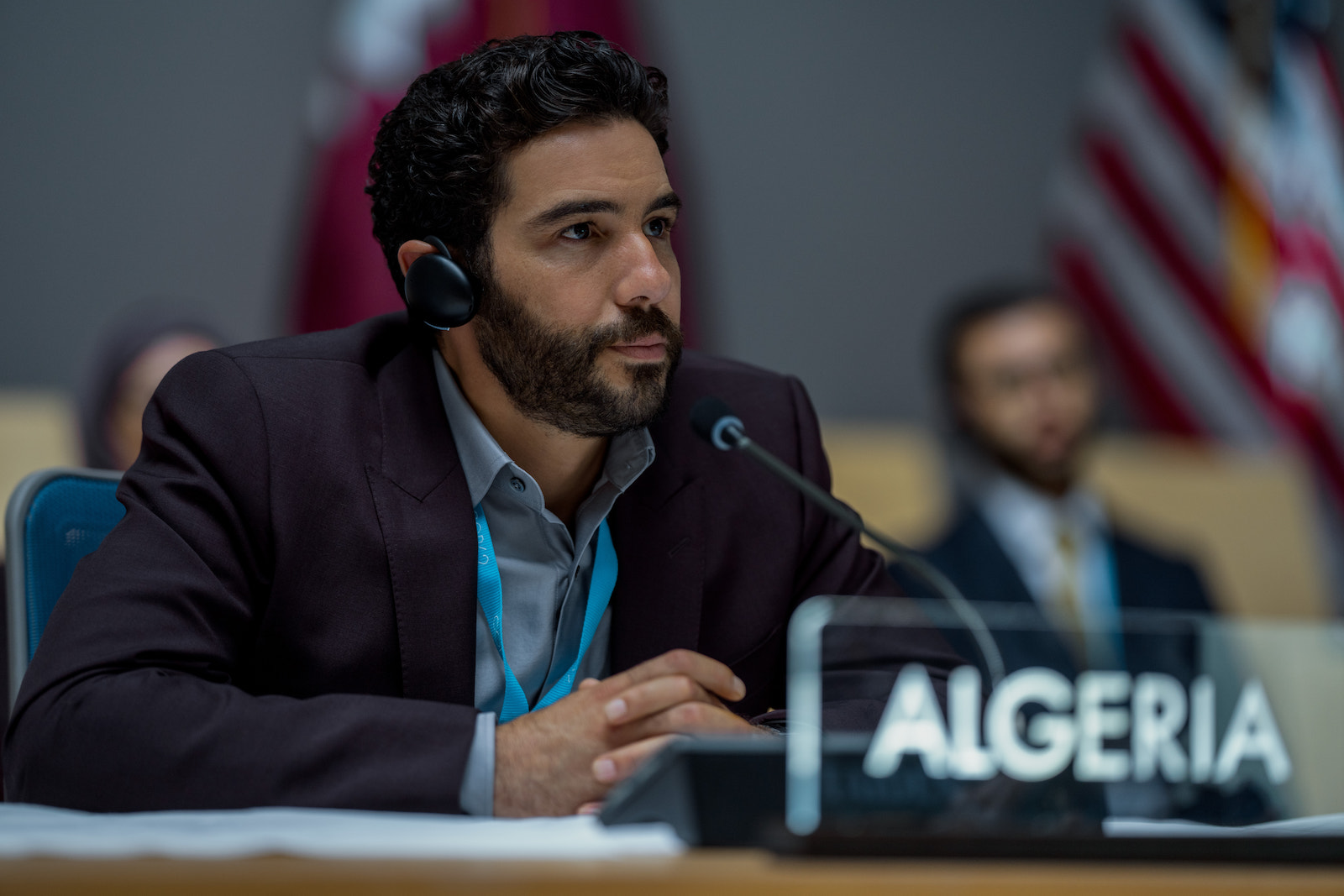 a man in a suit sits in front of an "Algeria" placard at a conference