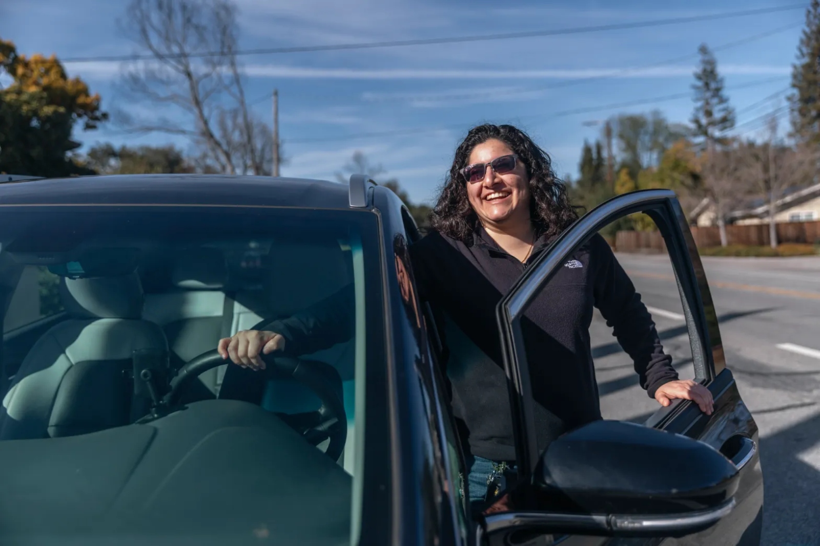 A smiling woman with curly black hair and sunglasses and a dark shirt stands outside the door of her black electric car.