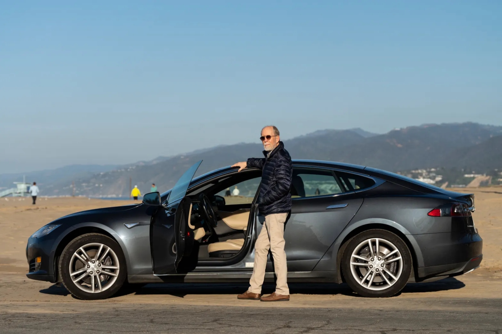 A balding man in loafters, khakis, and a down coat stands with his silver electric vehicle on the beach.