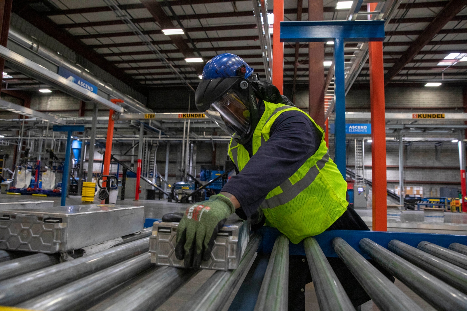 A man in a blue helmet and face shield with a neon yellow vest and green globes rolls a large metal box on a conveyor belt.