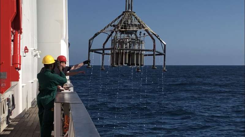 An octopus-like piece of equipment rises from the ocean next to a ship where two men stand, one taking a photo with a camera.