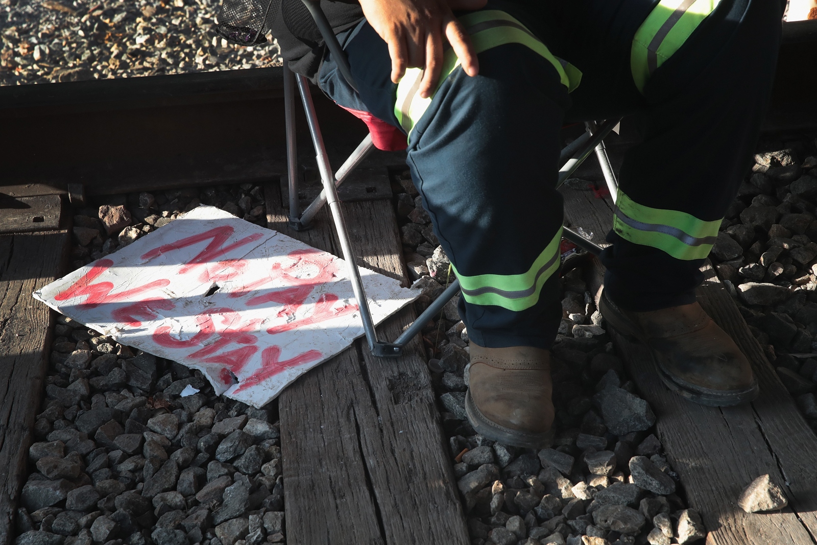 A man in black pants with yellow stripes along the bottom sits on a railroad track next to a handwritten sign that reads "No pay, we stay."