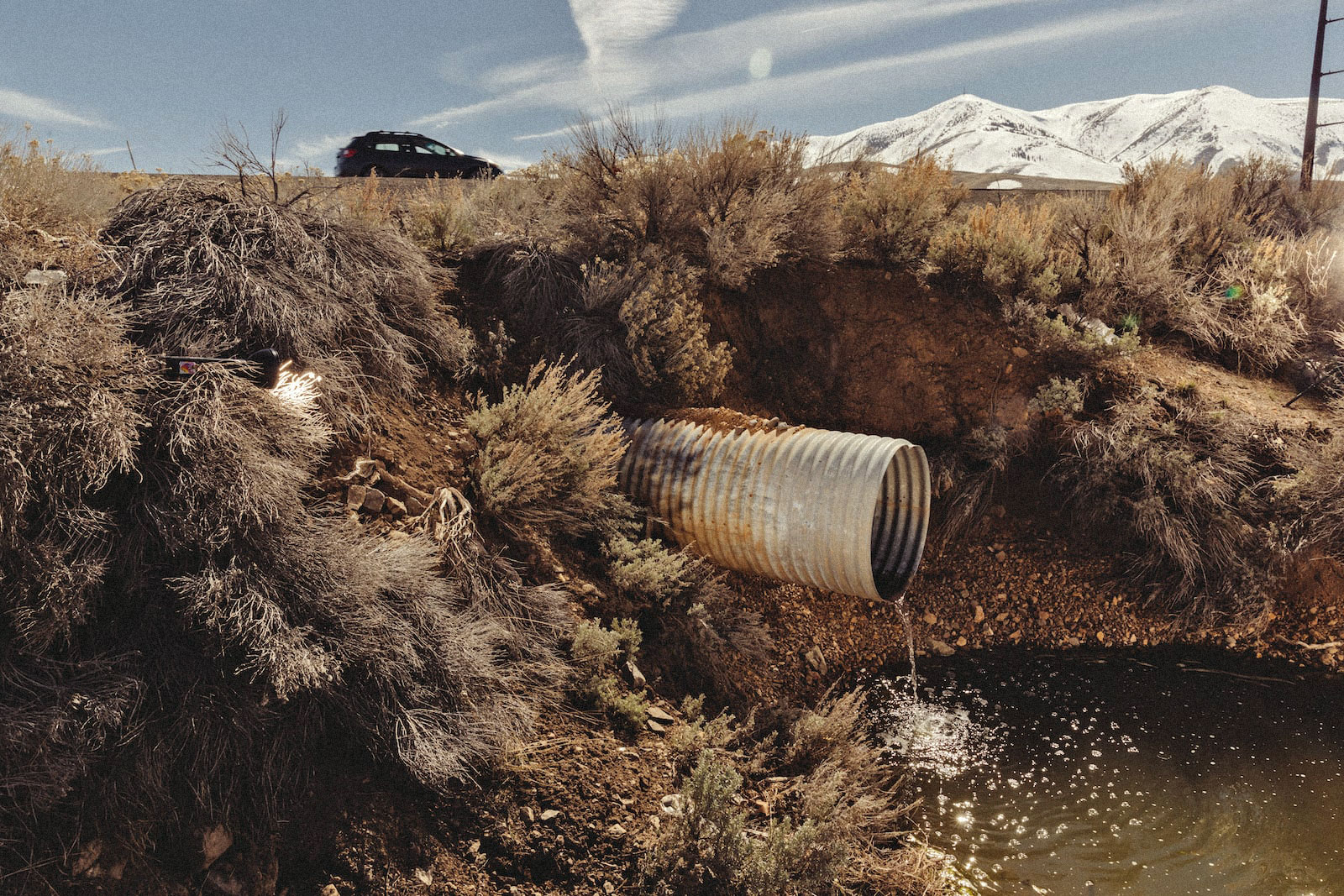 a drainpipe empties into a ditch while a car drives nearby