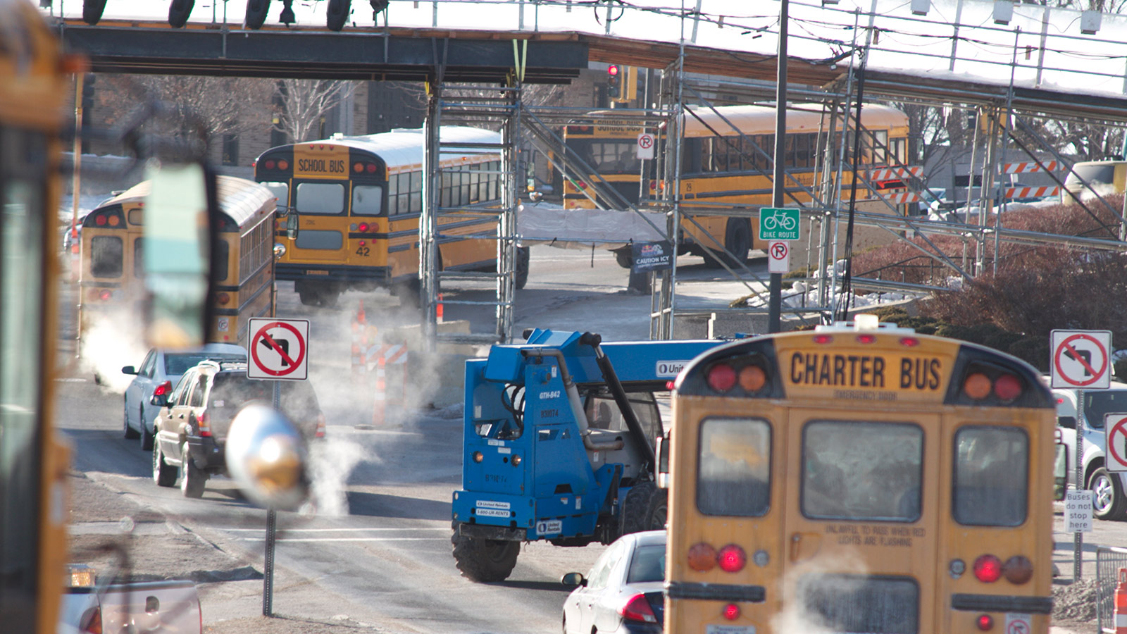 A row of yellow school buses drive down a street with exhaust coming from one of them.