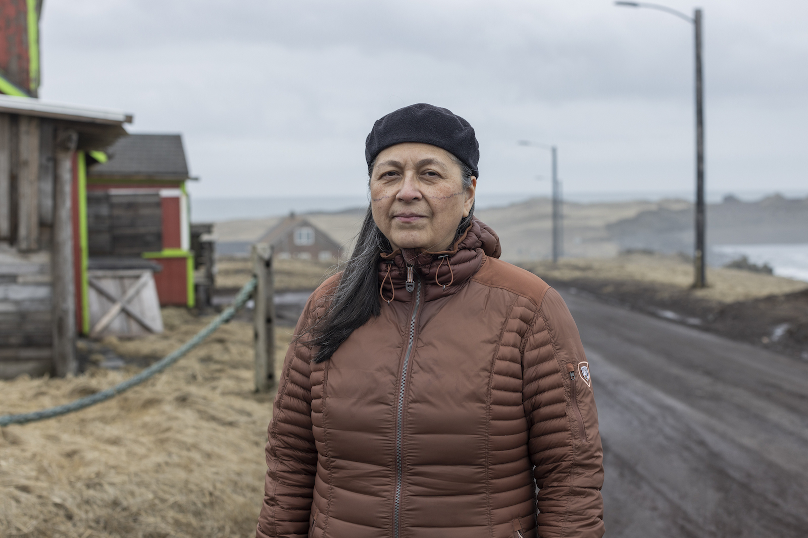 Historian Aquilina Lestenkof stands in a brown winter jacket and black hat, with the community of St. Paul behind her