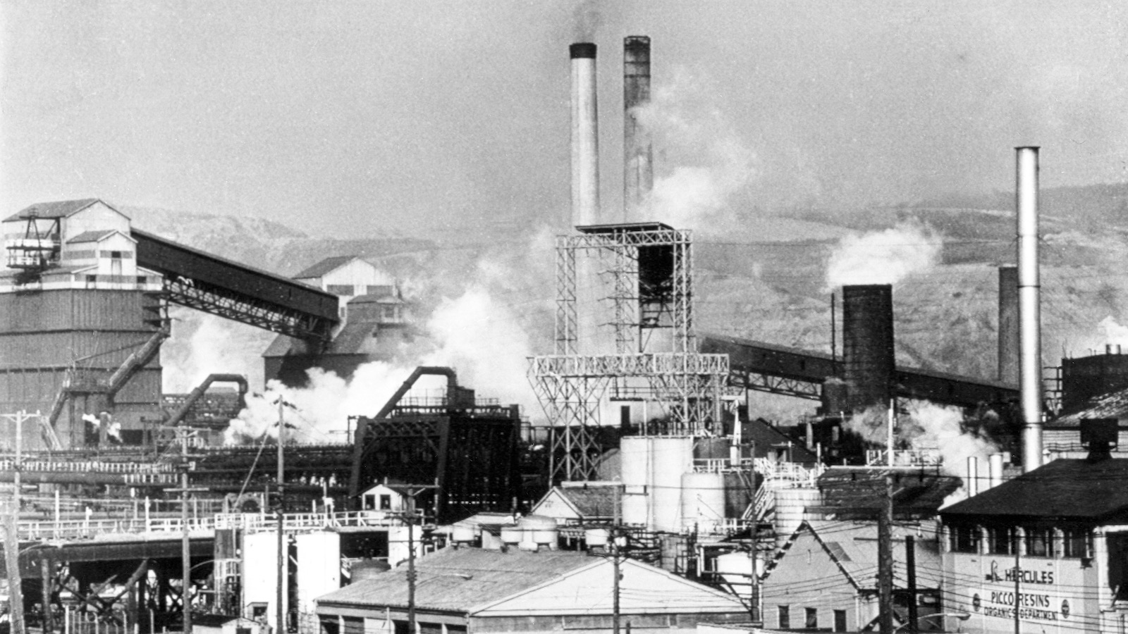 A coal coke factor spews smoke into the sky with a neighborhood in the foreground.