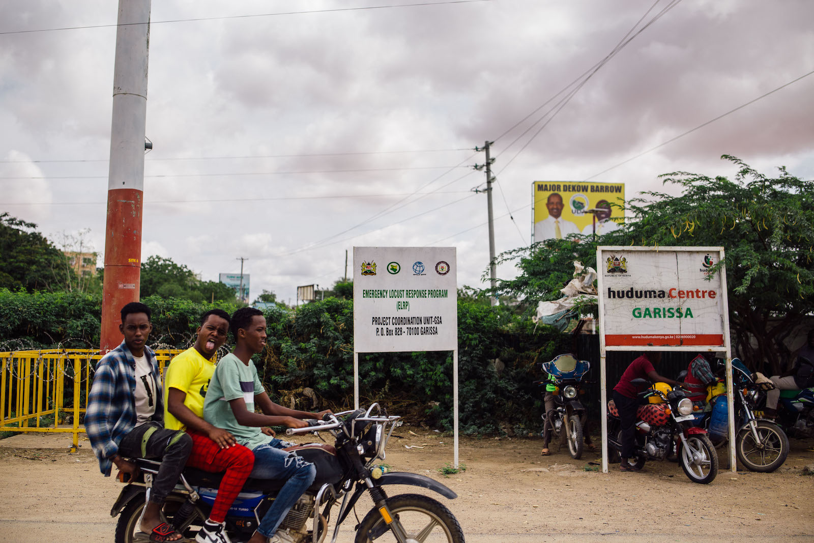 three young men ride on a motorcycle near two large signs