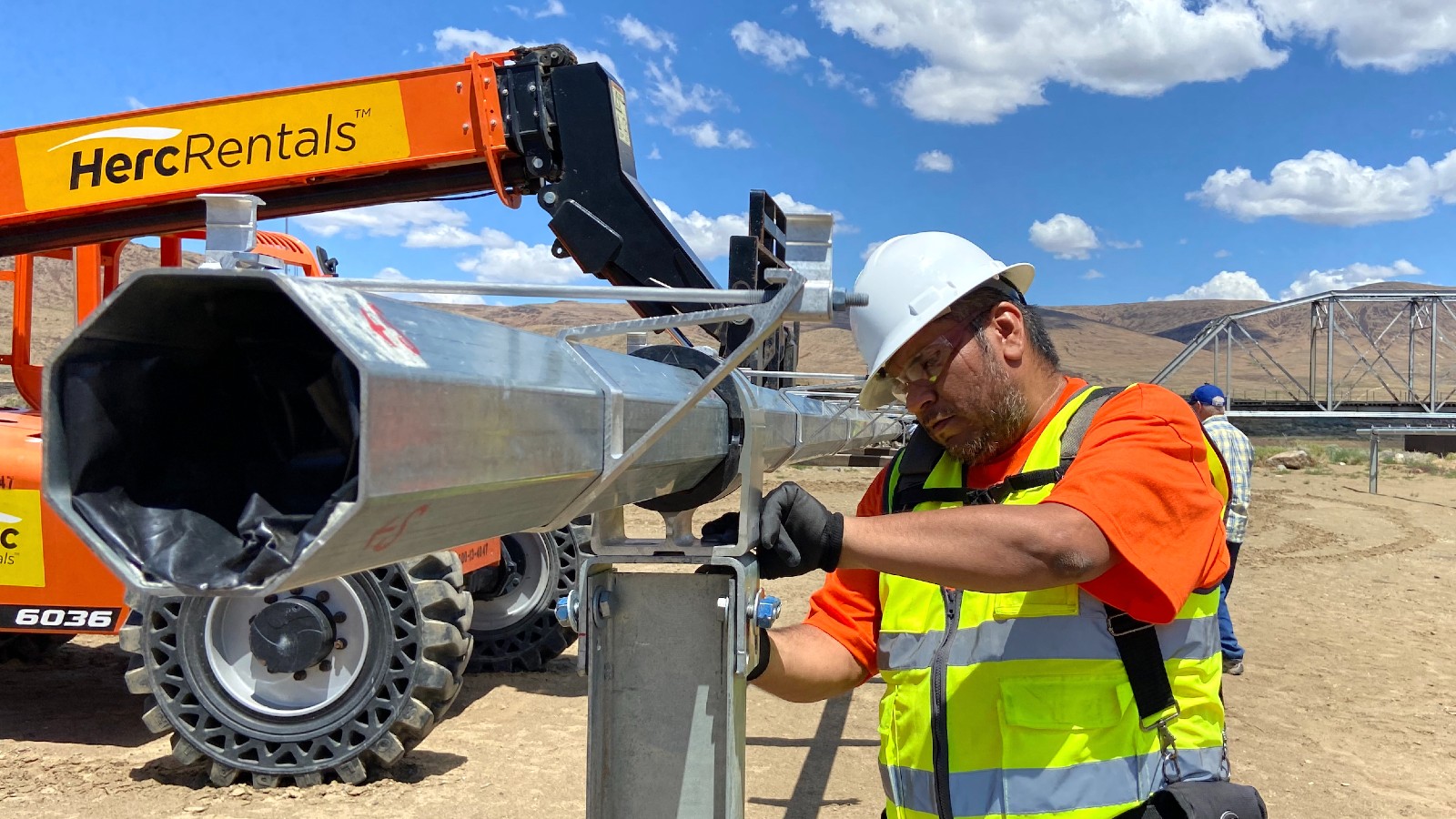 A man in a neon vest and white hard hat stands next to heavy machinery in the desert.