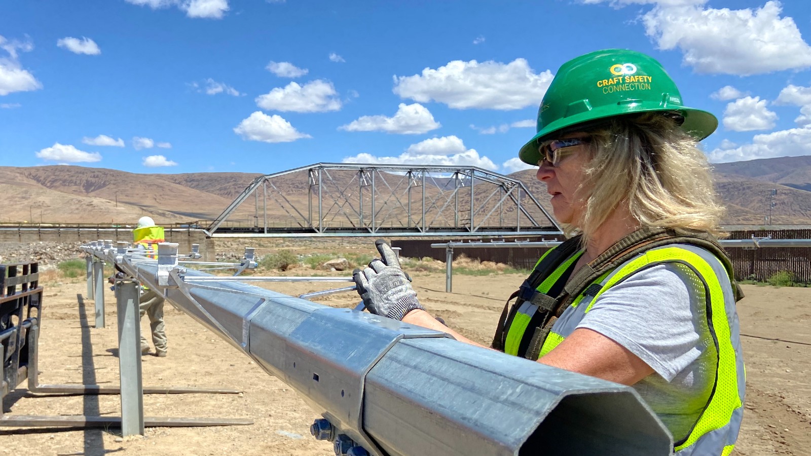 A woman in a green hard hat stands in front of a metal pole in the desert.