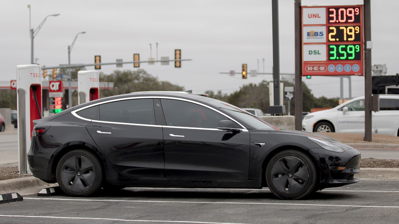 A black Tesla car charges in a parking lot next to a sign with gas prices.