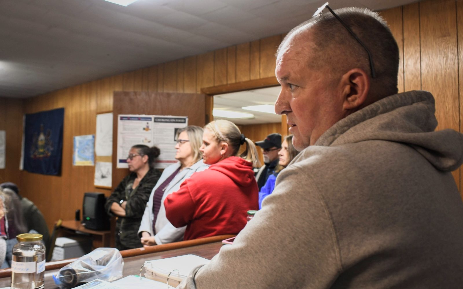 A group of people stand watching in a wood-paneled room.