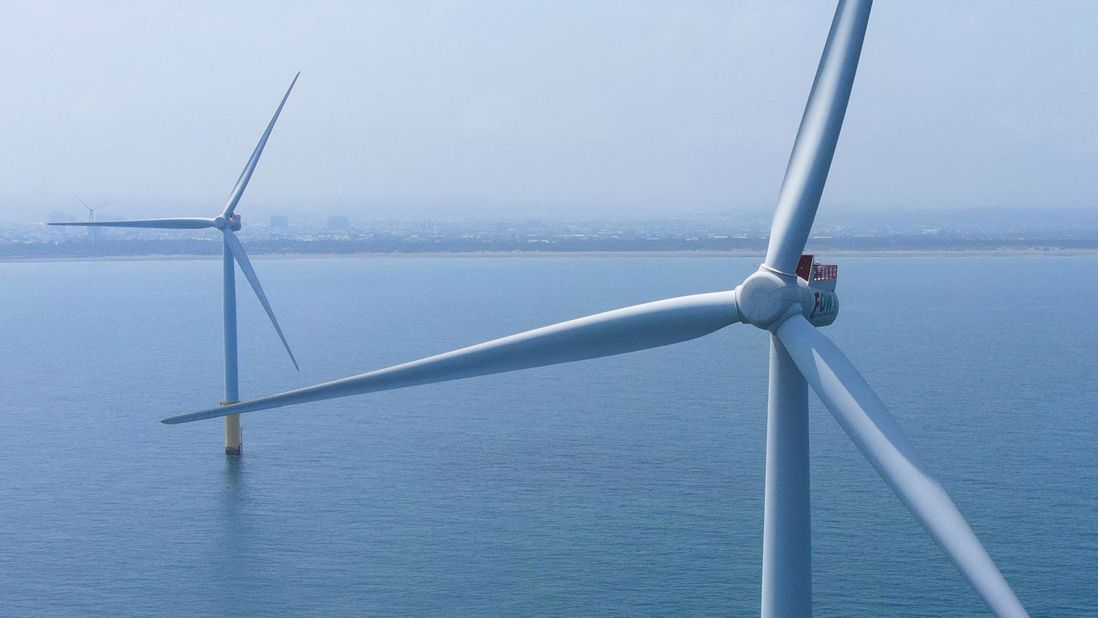 Aerial view of two wind turbines standing in sea water with shore in background