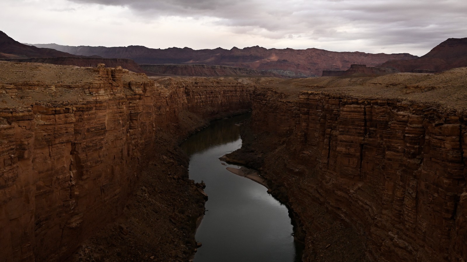 An aerial shot of a river cutting through red rocks under a cloudy sky.