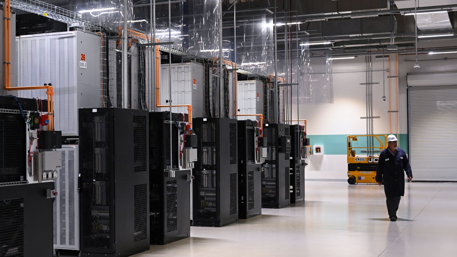 A man in a white hard hat and blue jumpsuit walks in an industrial space filled with large black boxes.