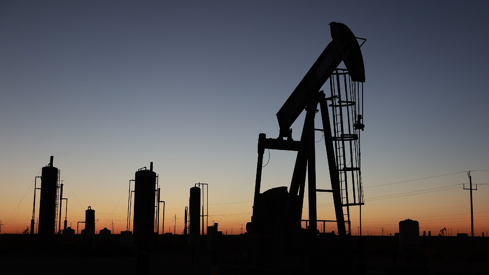 An oil rig and other industrial buildings stand against a sunset.