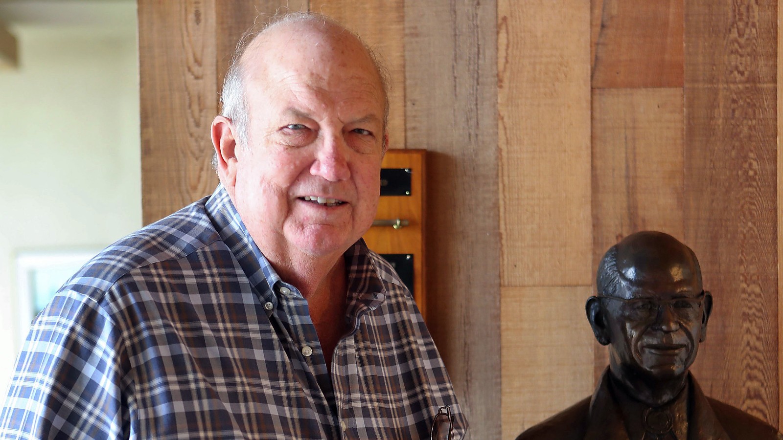 An older man in a plaid shirt stands next to a statue of a man.