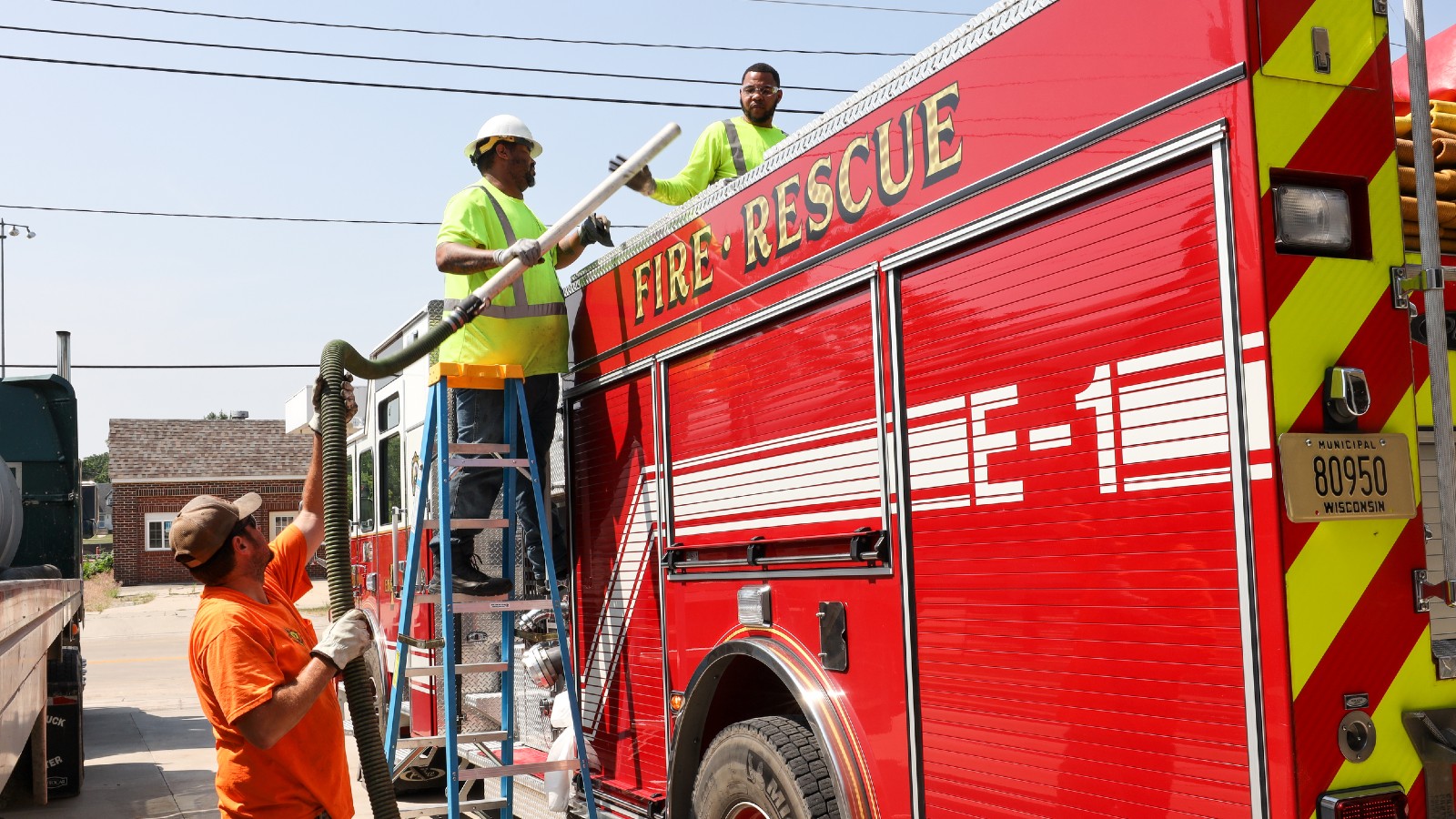 Two men manage a hos aboard a fire truck as another man looks on.