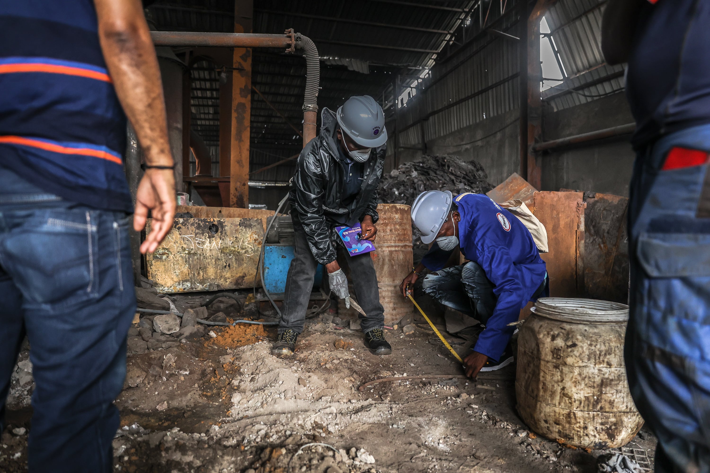 A man in a hard hat and jump suit, mask, and gloves stands next to a masked man as they reach for the ground.