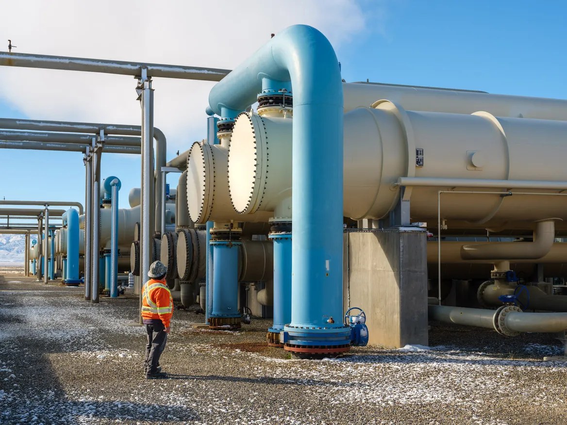 A man in a hard hat and an orange vest inspects large pipes.