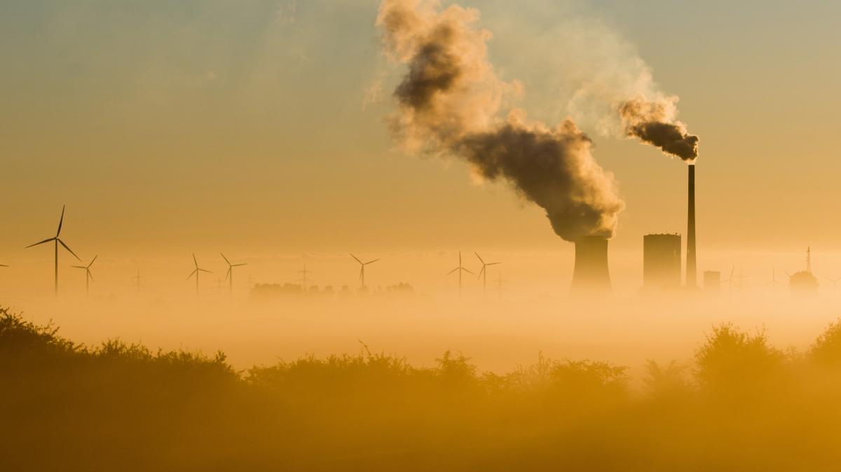 Haze settles over a valley where smoke rises from a nuclear plant and wind turbines spin in the distance.