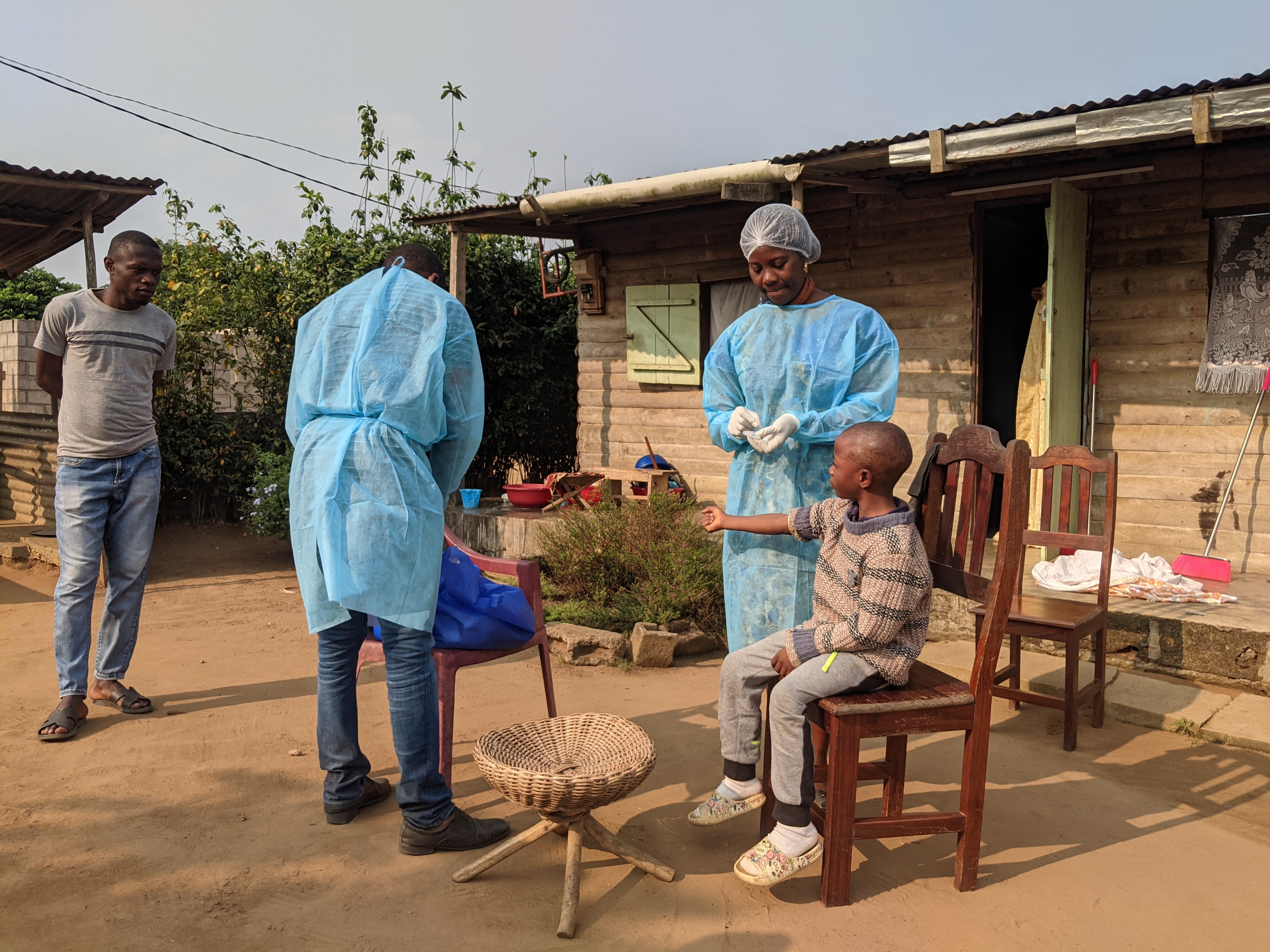 Two men in scrubs take blood from a boy sitting outside a house on a wooden chair.