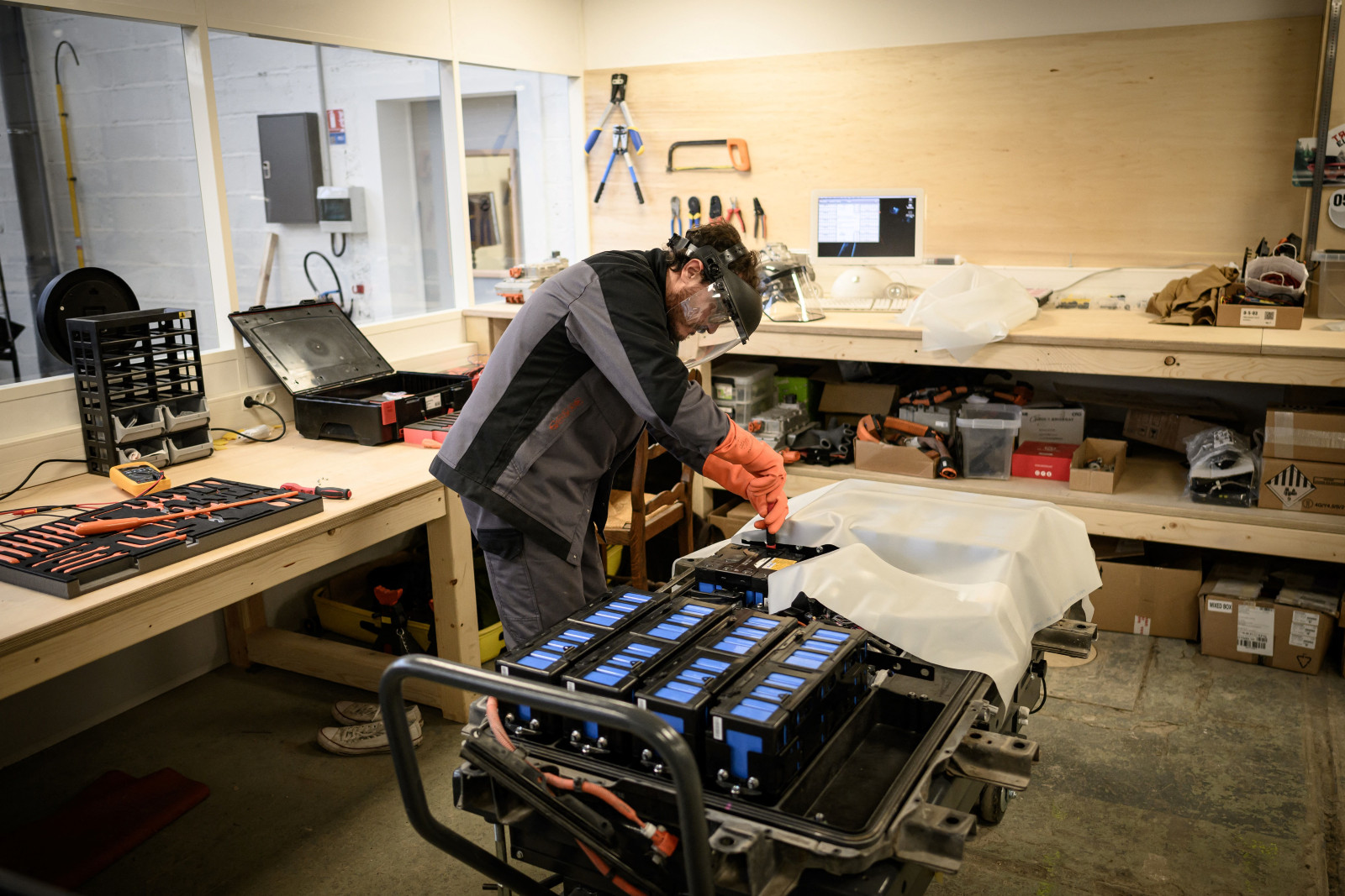 A man wearing a face shield, a gray jacket, and orange gloves leans over a large array of battery modules in a workshop