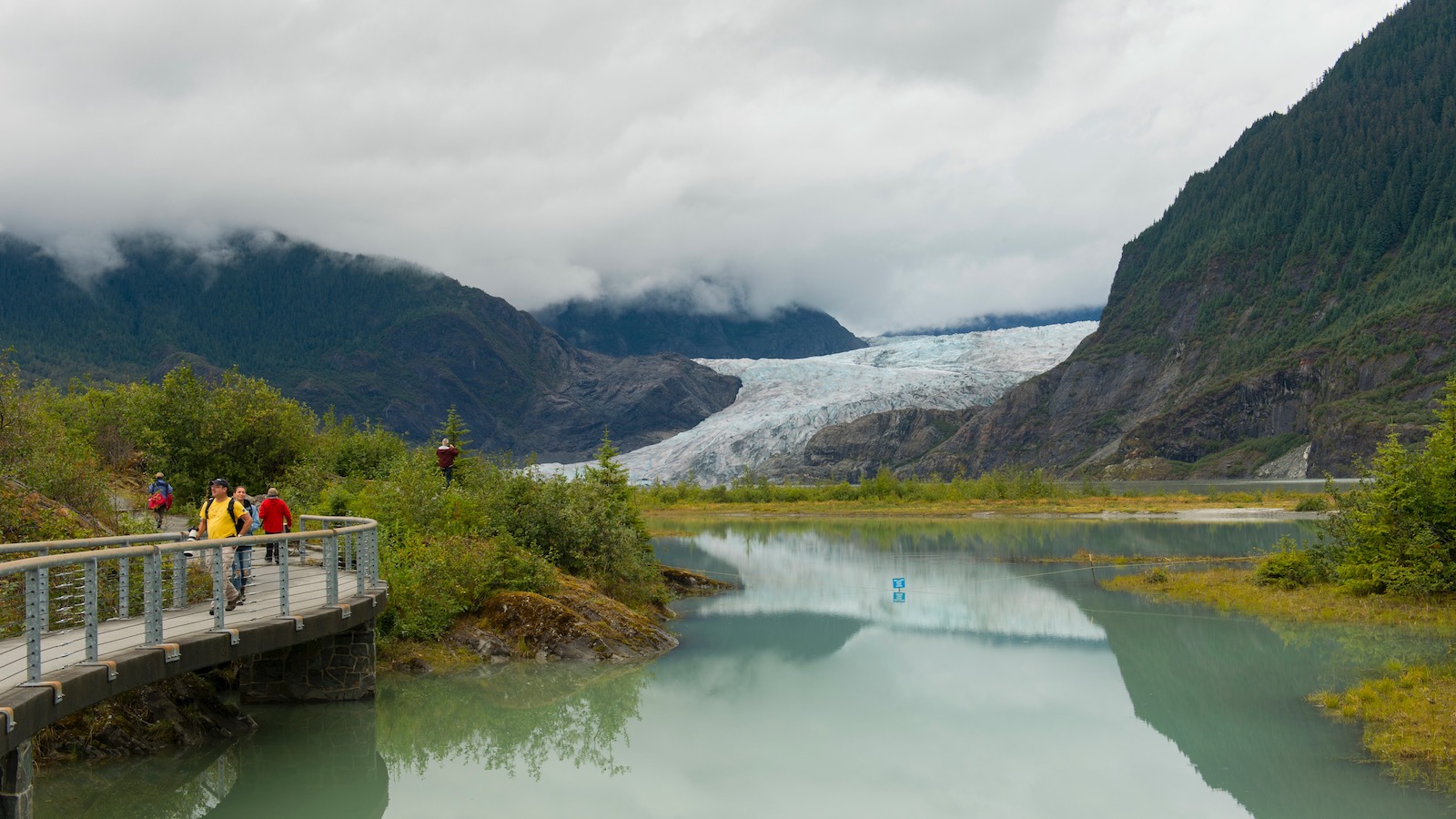 Four tourists walk along a boardwalk at Mendenhall Lake with Mendenhall Glacier in the background.